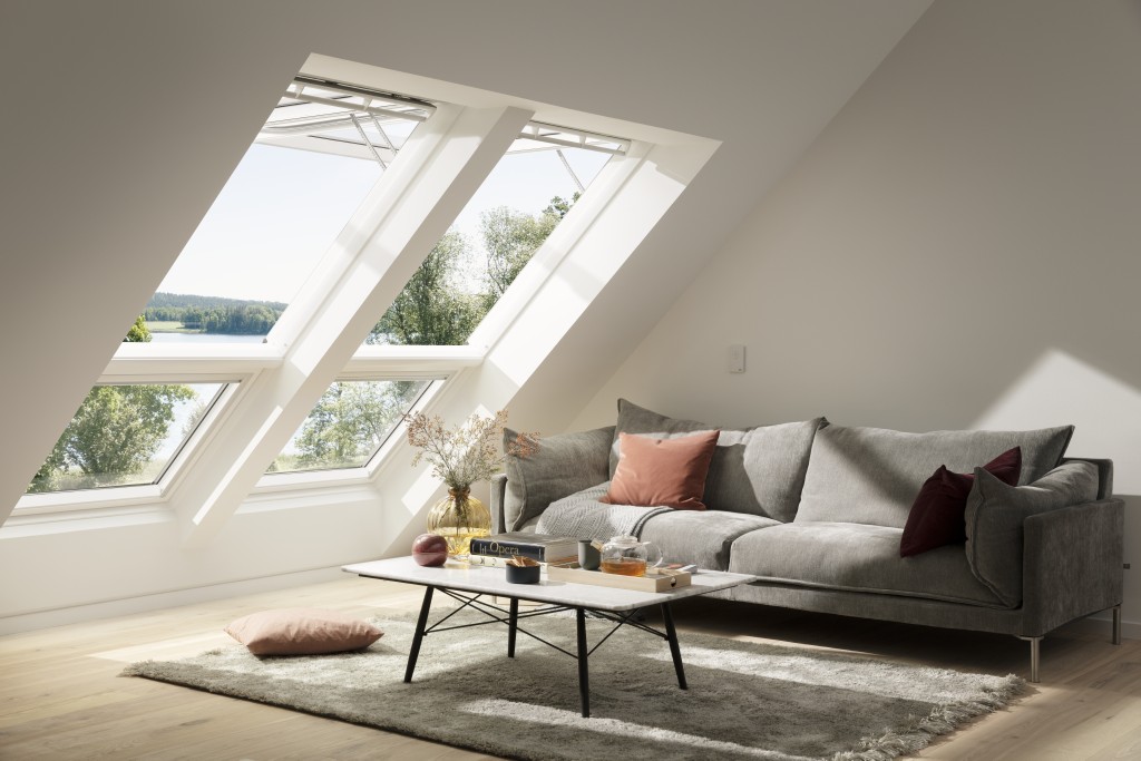 Wohnzimmer mit drei großen VELUX-Dachfenstern, grauem Sofa, Kissen, Teppich und niedrigem Tisch mit Dekoration.